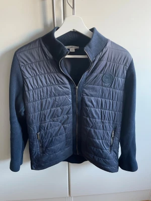 Mörkblå quiltad jacka Massimo Dutti - Snygg mörkblå quiltad jacka från Massimo Dutti med ribbad krage och ärmar. Jackan har två fickor med dragkedja och ett stilrent Massimo Dutti märke på bröstet. Materialmix med polyester och stickade detaljer ger en cool look.