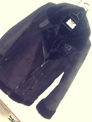 Svart pälsjacka med dragkedja - Säljer en svart pälsjacka från strandivarius med fluffig krage och muddar. Jackan har två snedställda fickor med dragkedja, detaljer med spänne vid halsen och en snygg asymmetrisk dragkedja framtill. Perfekt för dig som vill ha en cool och varm look.