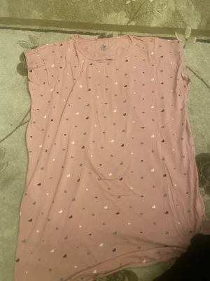 Rosa hjärtmönstrad topp ICA I Love Eco - Säljer en söt rosa topp från ICA I Love Eco i storlek L/XL. Toppen har ett gulligt hjärtmönster i olika färger och är tillverkad i mjuk ekologisk bomull. Modellen är ärmlös med rund halsringning, perfekt för varma dagar eller som mysplagg hemma.