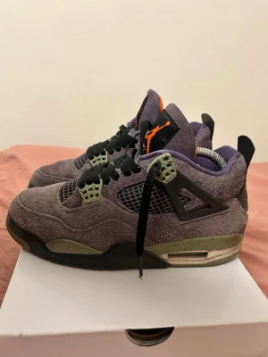 Nike Air Jordan 4 Retro lila mocka - Säljer ett par Nike Air Jordan 4 Retro i lila mocka med svarta och gröna detaljer. Skorna har svart snörning, meshpaneler och orange Jumpman-logga på plösen. Snygg kontrast mellan färgerna och klassisk siluett med robust sula. ( Box ingår 📦✅)