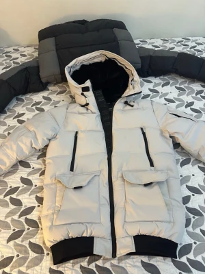 Beige pufferjacka Supply & Demand L - Snygg beige pufferjacka från Supply & Demand med svart fuskpäls i huvan och svarta detaljer. Jackan har flera fickor med dragkedja och lock, samt ribbade muddar. Perfekt för kalla dagar med sin tjocka och quiltade design. Det går att ta bort bälset om man vill