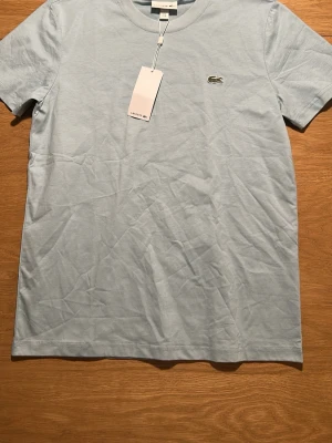 Ljusblå t-shirt från Lacoste - Snygg ljusblå t-shirt från Lacoste med klassisk krokodillogga broderad på bröstet. T-shirten har rund hals och korta ärmar, tillverkad i mjuk bomull som känns skön mot huden. Perfekt för dig som gillar stilrena och ikoniska plagg.