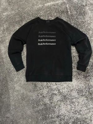 Svart Peak Performance sweatshirt - Svart sweatshirt från Peak Performance med tryckt logga i olika nyanser på bröstet. Tröjan har rund hals, ribbade muddar och är långärmad. Mjuk insida och klassisk passform, perfekt för chill eller träning.