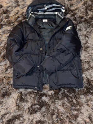 Svart pufferjacka från Burberry - Svart pufferjacka från Burberry med klassisk rutig huva i grått, vitt och svart. Jackan har dragkedja och knappar framtill, två sidofickor med dragkedja och långärmad passform. Perfekt för kalla dagar och har en schysst oversized vibe.