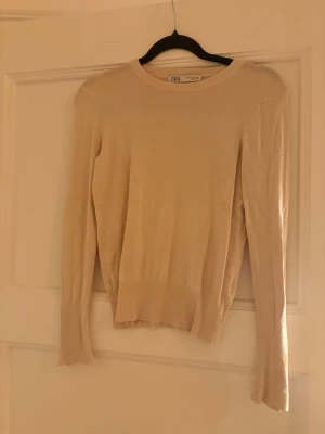 Beige långärmad tröja från Zara - Säljer en stilren beige långärmad tröja från Zara i tunn stickad kvalitet. Tröjan har rund halsringning och dekorativa knappar vid ärmsluten som ger en snygg detalj. Perfekt att matcha med jeans eller kjol för en clean look.