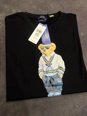 Svart Polo Bear t-shirt från Ralph Lauren - Svart t-shirt från Ralph Lauren med ikoniskt Polo Bear-tryck framtill. Klassisk rund hals och korta ärmar. Trycket visar en björn i preppy stil med RLTC-tröja och jeans. Skön bomullskvalitet och snygg passform, perfekt för en trendig look.