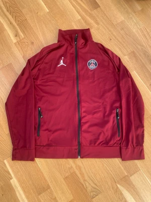 Vinröd PSG x Jordan zip-tröja - Snygg vinröd zip-tröja från Jordan och PSG med hög krage, svarta dragkedjor och loggor på bröstet. Perfekt för dig som gillar sportig stil och vill sticka ut med en ikonisk collab. Materialet är mjukt och lätt, med två fickor framtill.