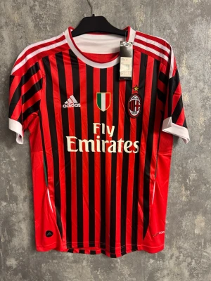 AC Milan Ibrahimovic matchtröja Adidas - Medium - Säljer en officiell AC Milan matchtröja från Adidas med Ibrahimovic och nummer 11 på ryggen. Tröjan har klassiska röda och svarta ränder, vita detaljer vid krage och ärmslut samt klubbmärke och sponsortryck på bröstet. Tillverkad i lätt polyester, perfekt för fotbollsälskare.