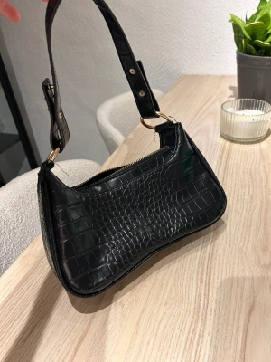 Väska Nelly - Svart handväska i skinn med guld-detaljer från Nelly