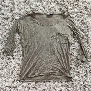 Beige långärmad tröja från Lindex  - En tunn och mjuk beige långärmad tröja från Lindex med rund halsringning och liten bröstficka på vänster bröst. Perfekt basic-plagg att ha i garderoben, lätt att matcha och skön att bära. Tillverkad i bomull för en luftig känsla. Tröjans är stretchig.  Färgen är mörk beige Storlek S 