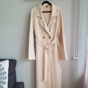 Trenchcoat - Beige trenchcoat som passar för lite varmare väder. Dubbelknäppning och knytskärp, inga fickor. Något liten i storleken och är enbart testad.
