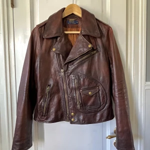 Ralph Lauren Läderjacka Nyskick - En ikonisk och tidlös läderjacka av Ralph Lauren. Skitsnygg och fantastisk kvalitet. Kragen kan bäras både platt och stående (rem under kragen). Många fickor, även innerficka. I princip aldrig använd då den tyvärr inte riktigt passade mig.