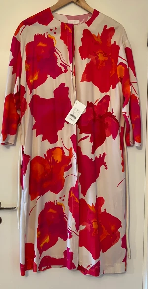 Carin wester - Helt nu klänning i beige med cerise och oranga blommor med dold knäppning och fickor man kan även ha den öppen som en kimono. Den är i lite kraftigare tyg och fint fall. Storlek 36 Nypris 1299kr