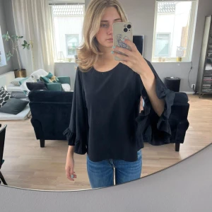 Svart topp  - Svart topp i ledig modell med volang på ärmen, knäpps med knapp i nacken. Ganska rymlig i storleken. 