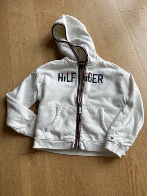 Tommy Hilfiger fodrad hoodie (barn 8-10) - Hoddie med Hilfiger över bröstet, med mjukt och lurvigt foder. Off-white med detaljer i blått och rött. Fickor och dragkedja. Bra kvalite och i fint skick. Använd under kort period men utan fläckar eller synligt slitage. Storlek M för barn, 8-10 år. 