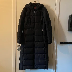 Áhkká svart dunkappa - Áhkká lång dunkappa med huva i nyskick. 2 fickor fram med dragkedja samt innerficka med dragkedja. Dragkedjor på sidorna längst ner för att vid behov göra kappan vidare. I ärmslut finns muddar i nylon. Mycket varm. Nypris 2999:-