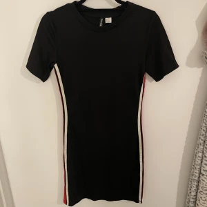 Klänning  - Svart klänning i storlek 42 med vita och röda ränder på sidorna. Sitter super snygg på och ger en snygg kroppsform av detaljerna på sidorna. Knappt använd! 💓