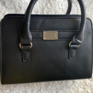 Svart handväska - Denna väska är från ur&penn. Har inte använt den på länge och tänkte sälja den. Väskan har en dragkedja, ett längre axelremsband men tyvärr går det inte att ta av. Väskan är rymlig, får plats med mobil, nycklar, Airpods och mer saker.