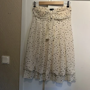 Klänning med hjärtan  - Vit/beige klänning med svarta hjärtan och volanger nedtill, vaddering vid brösten och inga axelband. Sitter jättefint på och går till ungefär mitten av låret (ref. Jag är 180cm). Nyskick, från ClubL. Storlek M/L. 