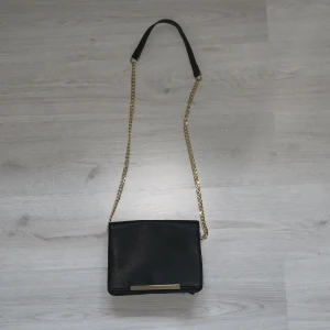 Svart handväska med guld detaljer  - Svart läder väska (fake) med guld detaljer. Den är i bra skick, vet inte var den är ifrån. Lagom storlek får plats med telfone, plånbok, nycklar mm