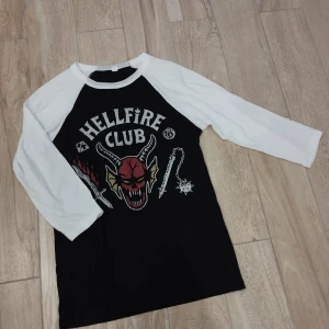 Hellfire Club Stranger Things Tröja  - Stranger Things tröja med 3/4 vita armar. Oversized till barn, passar till både vuxna och barn. Nypris var: ca 250 kr. Använd få gånger ca 2-4 ggr, alltså väldigt bra skick. Ny. 