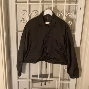 Kort, svart och tunn jacka – NY och OANVÄND - Kort svart jacka från H&M. Helt ny och oanvänd.  Storlek: M.  100 % polyester.  Hämtas i Vasastan, Stockholm. Eller skickas.
