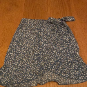 Blå blommig kjol - blå kjol med vita blommor, knut som man kan spänna midjan men den har en liten dragkedja som inte syns. 