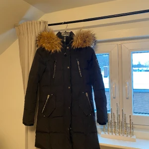 ROCKANDBLUE vinerjacka (Dam) (Svart) - En svart vinter jacka från Rockandblue. Luvan går lätt att ta av med en dragkedja och höjden går att justera med en spänntamp bak. Luvan är fodrad med päls, som går att ta av med knappar. Jackan har en kraftig tvåvägs dragkedja. På framsidan har den 5 fic