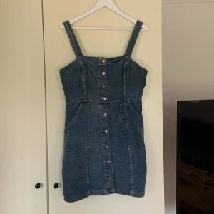 Jeansklänning - En jeansklänning med knappar hela vägen på framsidan, fickor på sidorna och justerbara axelband. Använd men i väldigt gott skick.