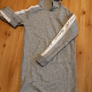 Oanvänd Gant sweatshirt klänning  - Grå sweatshirtklänning från Gant. Vita revärer med text längs ärmarna, ficka på magen samt luva.  