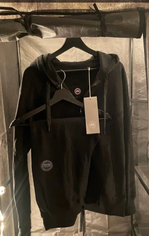 Canada goose dress  - Splitter ny SVART tracksuit från Canada Goose. Tillverkad av 100% bomull. Justerbar luva med dragsko, ribbstickad fåll och ärmslut och två handfickor framtill. Rund patch med logtoyp på vänster ärm. Samt Patch på vänsta sidan  På byxan 