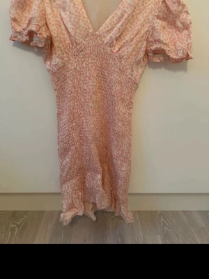 Ljusrosa kortklänning med volang - Söt ljusrosa kortklänning med små blommiga mönster, v-ringad hals och puffiga kortärmar. Klänningen har smock i midjan och volangkant nedtill för en feminin look. Perfekt för sommarens alla tillfällen.