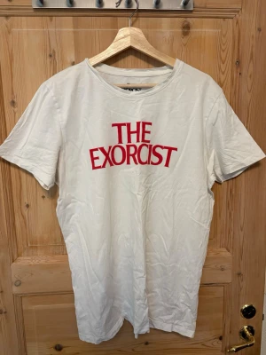 Vit The Exorcist t-shirt - Vit t-shirt med rött The Exorcist-tryck på framsidan och ett svartvitt motiv från filmen på baksidan. Klassisk rund hals och korta ärmar. Perfekt för dig som gillar skräckfilmer och vill ha en unik look.