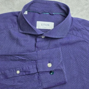Eton Chelsea skjorta lila storlek M / 39 / 15 1/2 - Lila skjorta från Eton i Slim-passform och modellen Chelsea. Diskret vävt ton-i-ton-mönster som ger skjortan ett elegant uttryck och passar lika bra till kostym som till chinos eller jeans. Tillverkad i 100% bomull med ett tunt och luftigt tyg som gör den särskilt lämpad för vår- och sommarsäsongen.  Storlek M / 39 Slim Fit  Mycket fint skick.  Mått Bröstbredd: ca 52 cm Plagglängd: ca 70 cm Axelbredd: ca 46 cm Ärmlängd: ca 64 cm  Material 100% bomull
