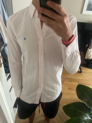 Ljusrosa randig skjorta med brodyr - Säljer en ljusrosa skjorta med tunna vita ränder och blå broderad detalj på bröstet. Skjortan har klassisk krage, knappar framtill och långa ärmar. Perfekt för dig som gillar stilrena och fräscha plagg.