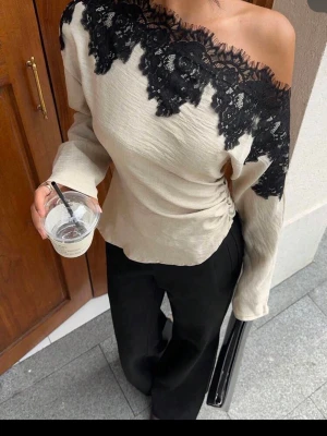 Beige offshoulder topp med spets - Trendig beige offshoulder topp med svart spetsdetalj längs halsringningen. Toppen har lång ärm och en lätt rynkning i sidan för en snygg passform. Perfekt för dig som vill sticka ut med en unik och elegant look.