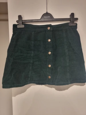 Grön manchesterkjol från Forever 21 - Mörkgrön kortkjol i manchester från Forever 21 med knappar framtill. Kjolen har en klassisk A-linjeform och är perfekt för dig som gillar retrostil. Snygg att matcha med stickade tröjor eller t-shirts.