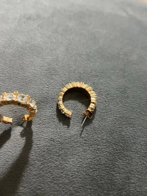 Guldiga creoler med pärldetaljer - Snygga örhängen i creolmodell med guldig finish och små vita pärlor längs kanten. Örhängena har en räfflad struktur som ger extra glans och en cool look. Perfekta för att lyfta din outfit med lite extra bling.