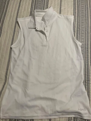 Vit ärmlös pikétopp från AONE - Snygg vit ärmlös pikétopp från AONE med klassisk krage och knappar framtill. Tillverkad i ett lätt och funktionellt material som andas, perfekt för sportiga aktiviteter. Enkel och stilren design med diskret logga nertill.