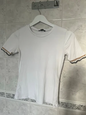 Vit t-shirt med spetsdetaljer - Vit t-shirt med korta ärmar och rund halsringning. Ärmsluten har fina spetsdetaljer i vitt, svart och en tunn guldfärgad kant. T-shirten är i mjuk och har en klassisk passform som funkar till det mesta💕