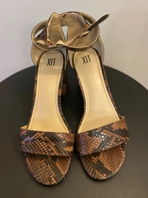 Bruna ormmönstrade pumps från XIT - Snygga pumps från XIT med brunt ormmönster, bred rem över tårna och justerbart ankelband med spänne. Skorna har en chunky klack och öppen tå. Materialet är konstläder med reptilstruktur för en trendig look.
