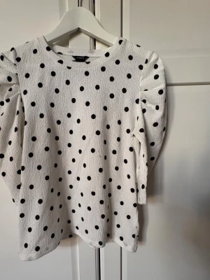 Vit prickig topp med puffärm - Säljer en vit topp med svarta prickar och puffärmar. Toppen har en rund halsringning och trekvartsärm. Materialet är lätt strukturerat och ger en cool look. Perfekt för dig som gillar trendiga detaljer och mönster.