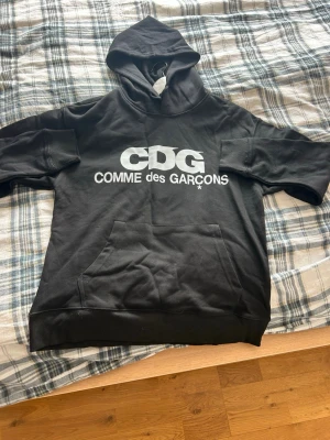 Svart hoodie Comme des Garçons - Svart hoodie från Comme des Garçons med stor vit CDG-logga på bröstet. Klassisk känguruficka framtill och huva. Tillverkad i mjukt material, perfekt för en avslappnad och trendig look.