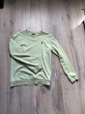 Ljusgrön sweatshirt från Lyle & Scott - Snygg ljusgrön sweatshirt från Lyle & Scott med klassisk rund halsringning och gul broderad logga på bröstet. Tröjan har ribbade muddar vid ärmslut och nederkant. Perfekt för en avslappnad och stilren look.