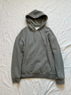 Grå hoodie från Pier One - Klassisk grå hoodie från Pier One med huva och dragsnören. Hoodien har en stor magficka och ribbade muddar vid ärmar och nederkant. Perfekt för en avslappnad och stilren look. Tillverkad i mjukt material för extra komfort.