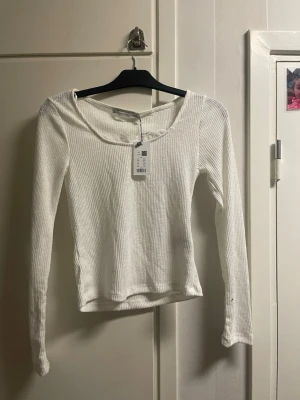 Vit ribbad långärmad topp - Säljer en vit ribbad långärmad topp med rund halsringning och diskret spetsdetalj vid halsen. Toppen har en slim passform och är tillverkad i ett mjukt, stretchigt material som passar perfekt till jeans eller kjol.