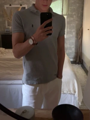 Grå pikétröja från Polo Ralph Lauren - Klassisk grå pikétröja från Polo Ralph Lauren i slim fit-modell. Tröjan har korta ärmar, krage och knappar framtill samt den ikoniska broderade loggan på bröstet. Tillverkad i mjuk bomull för en bekväm känsla.