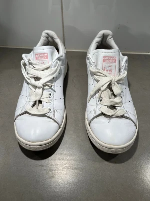 Adidas Stan Smith vita sneakers 38  - Klassiska vita Adidas Stan Smith sneakers med rosa detaljer på plösen. Skorna har snörning och är tillverkade i skinn med perforerade sidor för ventilation. Perfekta för en clean och stilren look.