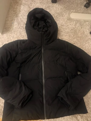Svart pufferjacka med huva Uniqlo - Snygg svart pufferjacka från Uniqlo. Perfekt till vintern. Har ett ganska litet hål över dem högra fickan som jag försökt sy ihop. Men man märker inte då de inte e så stort