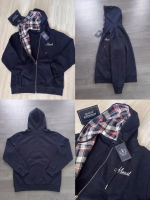 Svart hoodie med rutigt foder och dragkedja - Snygg svart hoodie med hel dragkedja och huva fodrad i röd, vit och svart rutigt tyg. Broderad text 'Marcel' på bröstet. Mjukt material och klassisk passform, perfekt för en chill och stilren look.
